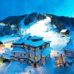 Verschneites Skigebiet in der Abenddämmerung mit beleuchteten Skipisten, mehreren Gebäuden und Kiefern, die das Gebiet umgeben.