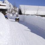 Zwei Personen fahren mit dem Schlitten einen verschneiten Weg zwischen hohen Schneebänken hinunter, mit Holzhäusern und Bergen im Hintergrund.