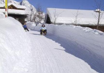 Zwei Personen fahren mit dem Schlitten einen verschneiten Weg zwischen hohen Schneebänken hinunter, mit Holzhäusern und Bergen im Hintergrund.