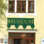 Ein gelbes Gebäude mit Holzfenstern und einem grünen Schild mit der Aufschrift "MUSEUM" über dem Eingang. Auf der linken Seite ist ein Baum zu sehen.