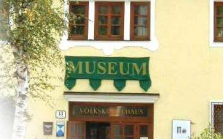 Ein gelbes Gebäude mit Holzfenstern und einem grünen Schild mit der Aufschrift "MUSEUM" über dem Eingang. Auf der linken Seite ist ein Baum zu sehen.