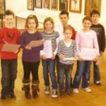 Sieben Kinder stehen in einer Kunstgalerie mit Papieren in der Hand und posieren für ein Gruppenfoto vor den an den Wänden ausgestellten Kunstwerken.