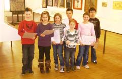 Sieben Kinder stehen in einer Kunstgalerie mit Papieren in der Hand und posieren für ein Gruppenfoto vor den an den Wänden ausgestellten Kunstwerken.