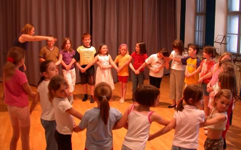 Eine Gruppe von Kindern steht in einem hell erleuchteten Raum im Kreis und hält sich an den Händen, während ein Erwachsener die Aktivität leitet.