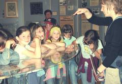 Eine Gruppe von Kindern steht um eine Vitrine herum, während ein Erwachsener auf sie zeigt und spricht, wahrscheinlich um eine Erklärung oder eine Führung in einem Museum oder einer Bildungseinrichtung zu geben.