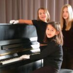 Drei Mädchen in schwarzer Kleidung stehen und sitzen an einem schwarzen Klavier vor grauen Vorhängen. Ein Mädchen sitzt am Klavier und trägt weiße Handschuhe.