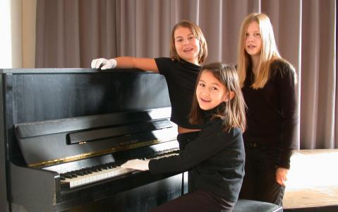 Drei Mädchen in schwarzer Kleidung stehen und sitzen an einem schwarzen Klavier vor grauen Vorhängen. Ein Mädchen sitzt am Klavier und trägt weiße Handschuhe.