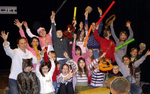 Eine Gruppe von Kindern und Erwachsenen posiert in einem Innenraum, hebt die Arme und hält Musikinstrumente in der Hand, wirkt begeistert und feierlich.