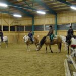 Vier Personen reiten auf Pferden in einer Reithalle mit Sandboden und Holzwänden.