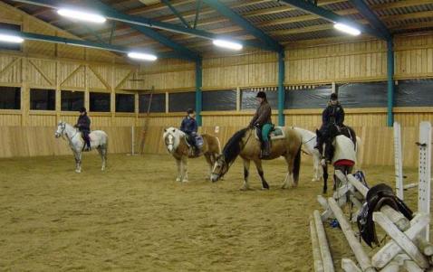 Vier Personen reiten auf Pferden in einer Reithalle mit Sandboden und Holzwänden.