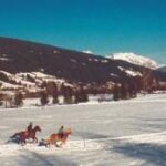 Fünf Personen reiten auf Pferden über ein verschneites Feld mit Bergen und Bäumen im Hintergrund unter einem klaren blauen Himmel.