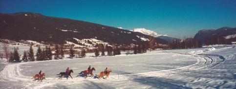 Fünf Personen reiten auf Pferden über ein verschneites Feld mit Bergen und Bäumen im Hintergrund unter einem klaren blauen Himmel.