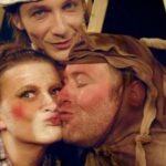 Drei Erwachsene in theatralischen Tierkostümen posieren eng beieinander; einer küsst den anderen auf die Wange, während der dritte im Hintergrund lächelt.