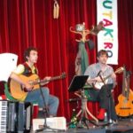 Zwei Musiker sitzen auf der Bühne und spielen Gitarre und Banjo, umgeben von Musikinstrumenten und Requisiten, vor roten Vorhängen und einem vertikalen Banner mit der Aufschrift "UTAKIRO".