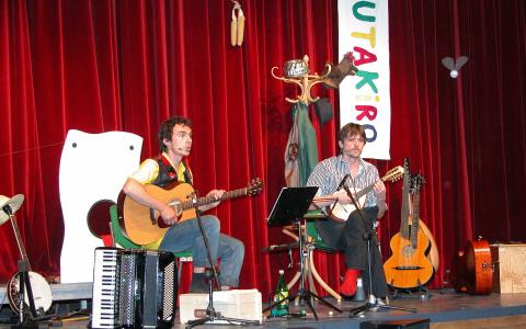 Zwei Musiker sitzen auf der Bühne und spielen Gitarre und Banjo, umgeben von Musikinstrumenten und Requisiten, vor roten Vorhängen und einem vertikalen Banner mit der Aufschrift "UTAKIRO".