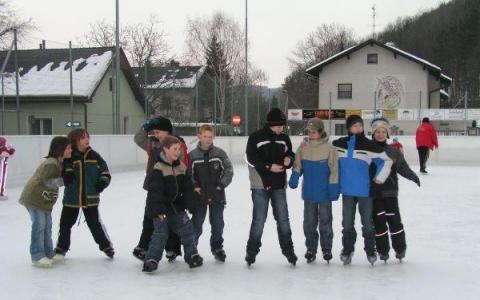 Eine Gruppe von Kindern in Winterkleidung steht zusammen auf einer Eislaufbahn im Freien mit Häusern und Bäumen im Hintergrund.