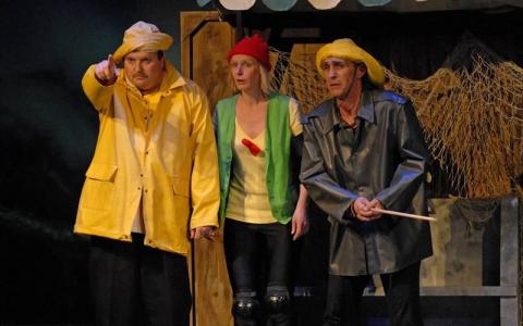 Drei Personen in bunten Kostümen, darunter gelbe Regenmäntel und Hüte, stehen auf der Bühne vor einer rustikalen Kulisse mit Fischernetzen. Eine Person deutet nach vorne.