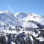 Die schneebedeckten Gebäude des Skigebiets schmiegen sich am Fuße eines Berghangs unter einem strahlend blauen Himmel an die Pinienbäume.