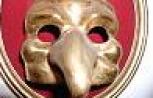 Eine goldfarbene Maske mit einer langen, schnabelartigen Nase und runden Augenlöchern auf rotem Hintergrund.