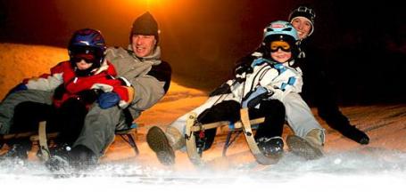 Zwei Erwachsene und zwei Kinder fahren nachts in Winterkleidung und mit Helmen auf Holzschlitten einen verschneiten Hügel hinunter.