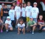 Eine Gruppe von Kindern posiert vor einem Feuerwehrauto, einige stehen, andere hocken, alle tragen Freizeitkleidung und lächeln.