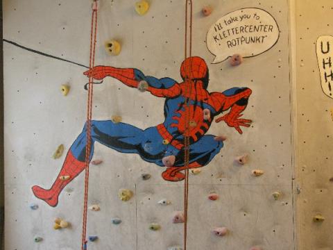 Eine Kletterwand mit einem Bild von Spider-Man und einer Sprechblase mit der Aufschrift "Ich bringe dich zum Klettercenter Rotpunkt". An der Wand sind Griffe zum Klettern angebracht.