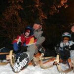 Vier Personen in Winterkleidung, die nachts auf Schlitten im Schnee sitzen und lächeln, mit einer Straßenlaterne und Bäumen im Hintergrund.