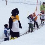 Eine Gruppe kleiner Kinder in Skikleidung steht in der Schlange auf einem schneebedeckten Förderband neben einer Person im Pinguinkostüm und einem erwachsenen Betreuer.