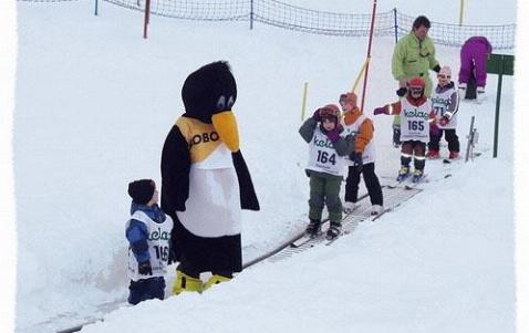 Eine Gruppe kleiner Kinder in Skikleidung steht in der Schlange auf einem schneebedeckten Förderband neben einer Person im Pinguinkostüm und einem erwachsenen Betreuer.
