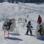 Ein Kind fährt auf Skiern durch einen verschneiten Parcours, der mit Cartoon-Pinguin-Schildern markiert ist, und wird von einem Erwachsenen in Rot beaufsichtigt.