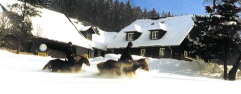 Zwei Menschen reiten auf Pferden durch den Schnee vor einem verschneiten Haus, mit Bäumen und einem Wald im Hintergrund unter blauem Himmel.