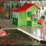 Zwei kleine Kinder mit Schwimmflügeln spielen in der Nähe eines Hallenbads; eines ist im Wasser, das andere steht daneben, mit einem bunten Spielhaus und Spielzeug im Hintergrund.