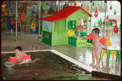Zwei kleine Kinder mit Schwimmflügeln spielen in der Nähe eines Hallenbads; eines ist im Wasser, das andere steht daneben, mit einem bunten Spielhaus und Spielzeug im Hintergrund.