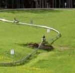 Zwei Personen fahren mit Ein-Personen-Karts eine kurvenreiche alpine Rutsche auf einem von Bäumen gesäumten Grashügel hinunter.