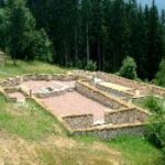 Ruinen alter Steinbauten, umgeben von Gras und Bäumen, mit einem Waldgebiet im Hintergrund.