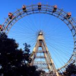 Großes Riesenrad mit roten Fahrgastkabinen vor einem klaren blauen Himmel, teilweise verdeckt durch Bäume im Vordergrund.