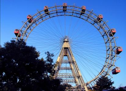 Großes Riesenrad mit roten Fahrgastkabinen vor einem klaren blauen Himmel, teilweise verdeckt durch Bäume im Vordergrund.