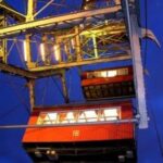 Zwei rot beleuchtete und mit Nummern versehene Riesenradkabinen, von unten gesehen vor einem tiefblauen Abendhimmel.