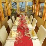 Ein langer Esstisch mit weißen Stühlen, Servietten, Gläsern und einem roten Tischläufer in einem sonnendurchfluteten Raum mit großen Fenstern und Stadtblick.