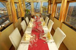 Ein langer Esstisch mit weißen Stühlen, Servietten, Gläsern und einem roten Tischläufer in einem sonnendurchfluteten Raum mit großen Fenstern und Stadtblick.