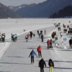 Auf einem großen zugefrorenen See, der von schneebedeckten Bergen umgeben ist, gehen die Menschen spazieren und laufen Schlittschuh, und entlang des Eises sind Gruppen versammelt und Schilder aufgestellt.