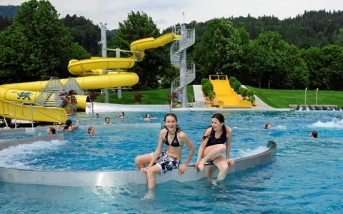 Zwei Mädchen sitzen am Rande eines öffentlichen Freibads mit einer gelben Wasserrutsche und anderen Badegästen im Hintergrund, umgeben von Bäumen und Grünflächen.