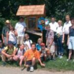Eine Gruppe von Menschen, darunter Kinder und Erwachsene, posieren gemeinsam im Freien vor einem kleinen Holzbau mit Schindeldach.