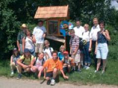 Eine Gruppe von Menschen, darunter Kinder und Erwachsene, posieren gemeinsam im Freien vor einem kleinen Holzbau mit Schindeldach.