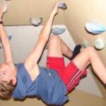 Ein kleiner Junge in Sportkleidung klettert an einer Kletterwand, hält sich an den Griffen fest und stützt sich mit den Füßen ab.