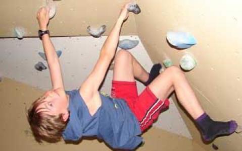 Ein kleiner Junge in Sportkleidung klettert an einer Kletterwand, hält sich an den Griffen fest und stützt sich mit den Füßen ab.