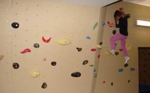 Ein Kind in einem schwarzen Hemd und einer rosa Hose klettert an einer Boulderwand mit bunten Griffen.