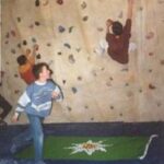 Drei Kinder klettern an einer Indoor-Kletterwand über blaue Matten, auf dem Boden liegt eine grüne Matte mit einer weißen Blume.