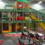 Indoor-Spielplatz mit Klettergerüsten, Rutschen, Softplay-Bereich und mehreren Tret-Go-Karts; ein Erwachsener sitzt in der Nähe.