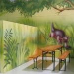 Holztische und -bänke stehen zwischen Wänden, die mit einem Dschungelthema bemalt sind, darunter Grünpflanzen und die Abbildung eines Dinosauriers.